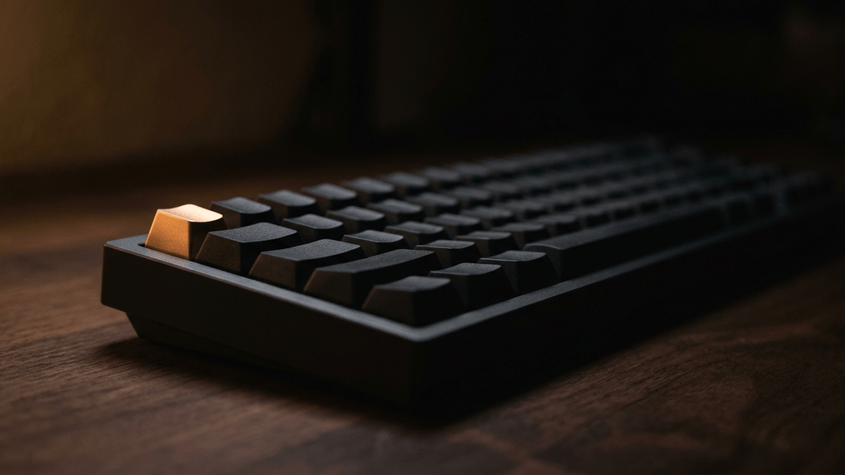 Illustration: Nahaufnahme einer dunklen mechanischen Tastatur im Halbdunkel, die Tastenkappen sind vollständig blank und unbeschriftet, eine einzelne Taste am linken Rand der oberen Reihe fängt warmes Licht