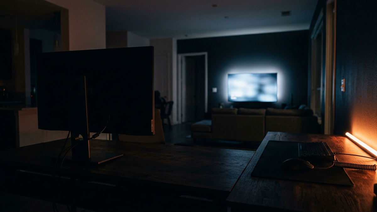 Blick über einen Schreibtisch mit Monitor auf einen leuchtenden Fernseher am anderen Ende des dunklen Raums