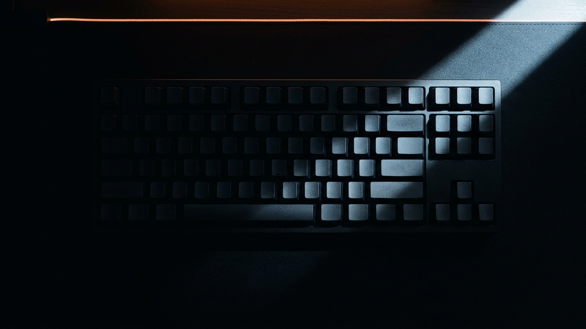 Eine schwarze Tastatur von oben, fast ganz im Schatten, nur ein schmales Lichtband quert die unbeschrifteten Tasten