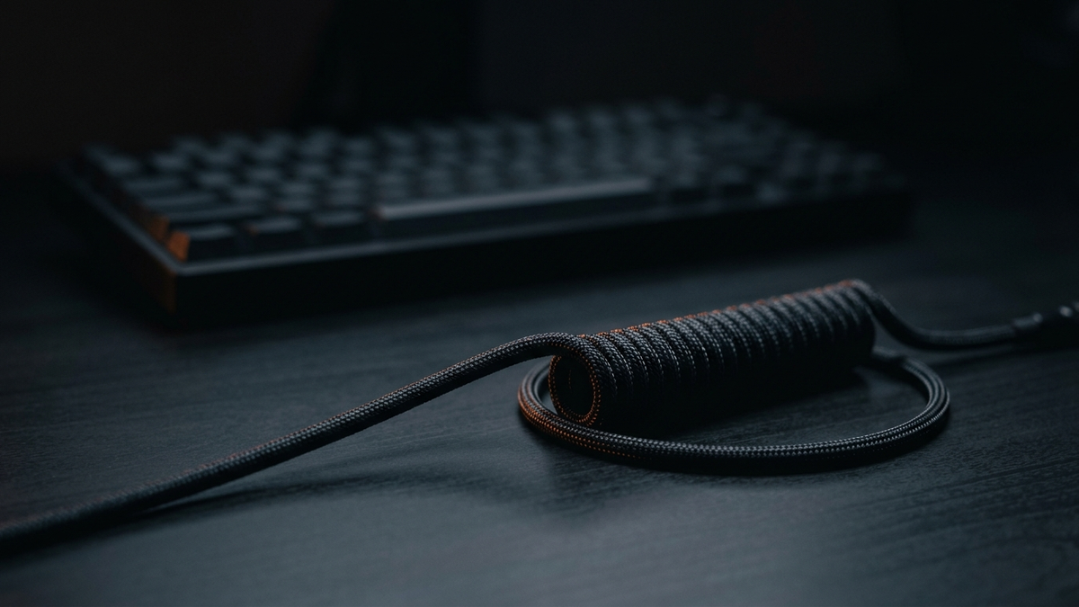 Ein gewendeltes Kabel liegt scharf im Vordergrund, dahinter unscharf die Silhouette einer Tastatur